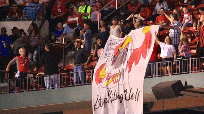 Protesters unfurled a banner of a Cardinals mascot wearing a jersey that says "Expect Us" and holding a "Racism Lives Here" sign.
