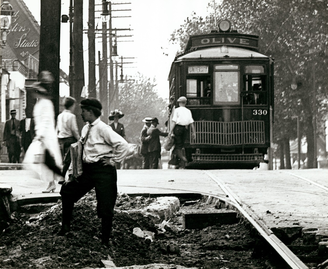 A newsboy stands in the rubble of street repair work on Olive Boulevard looking west toward Grand Avenue, ca. 1907. In the background, the Olive Boulevard streetcar line stops to pick up passengers near the photographic studio of Emme and Mayme Gerhard (visible on the left hand side of the image).