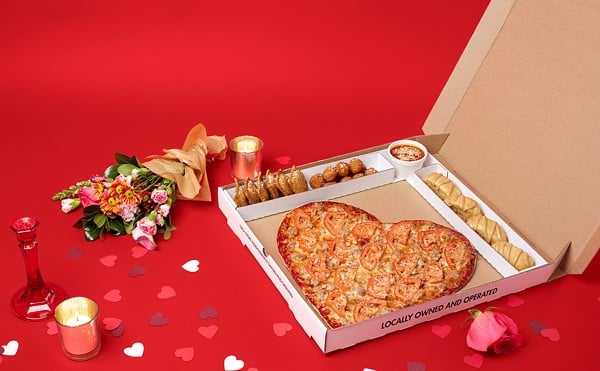 Heart-shaped pizza is in a box with a candle and flowers next to it.