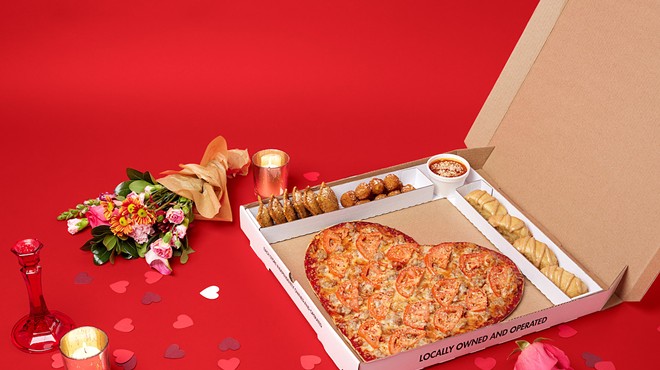 Heart-shaped pizza is in a box with a candle and flowers next to it.