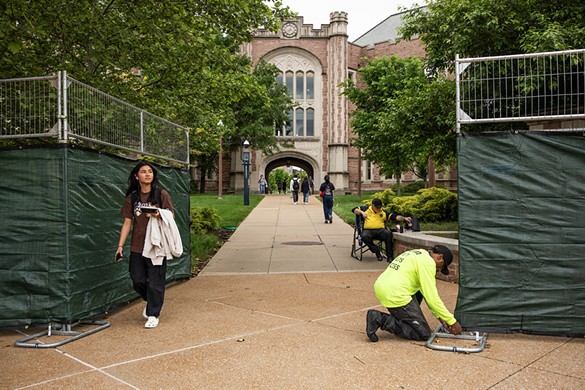 Students walk past workers installing a green tarp along the metal fence that surrounds Wash U’s Danforth Campus on Monday, May 6, 2024.