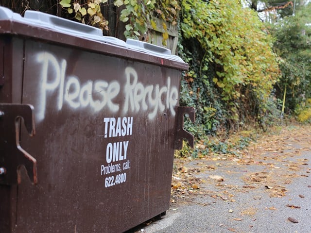 The words "Please Recycle" are spray painted above a brown dumpster that reads "Trash Only."