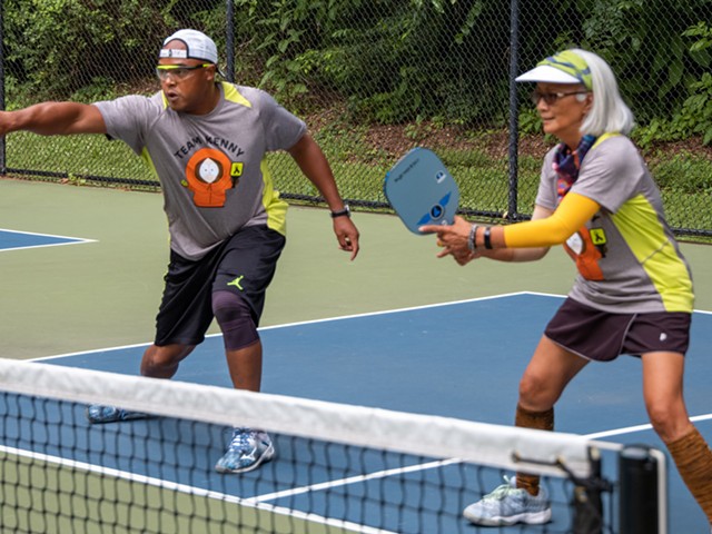 Two people stand with rackets in front of a net on a pickle-ball court.