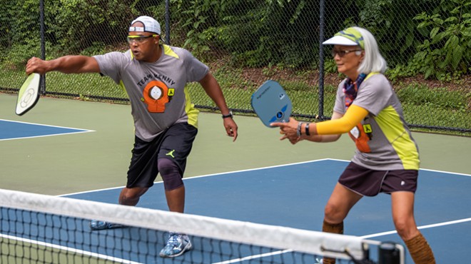 Two people stand with rackets in front of a net on a pickle-ball court.