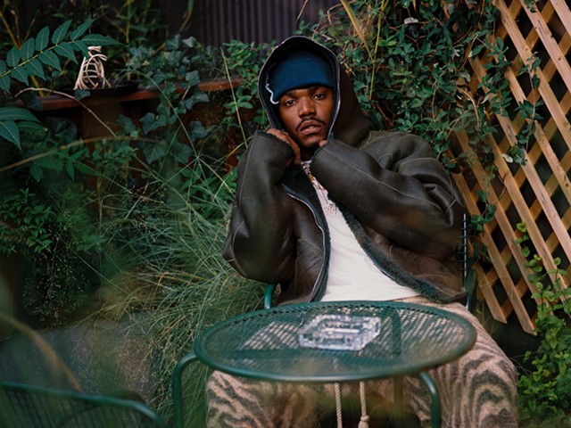 Smino pulls a hoodie over his head as he sits in a chair in a yard.