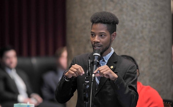 State Representative Rasheen Aldridge, wearing a suit jacket, button-down shirt and a tie, speaks in front of a microphone.
