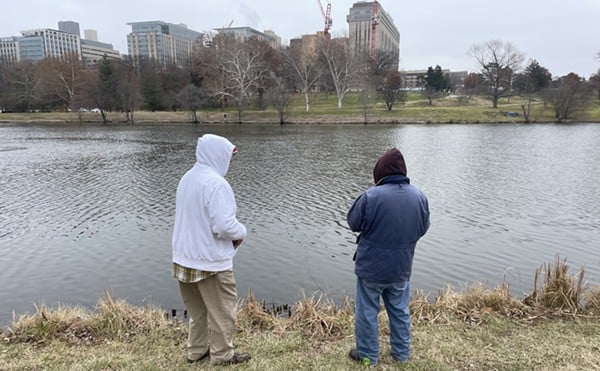 Two men stand in front of a lake in Forest Park with their backs turned to the camera. The man to the left wears a white hoodie and the man to the left wears a blue jacket.