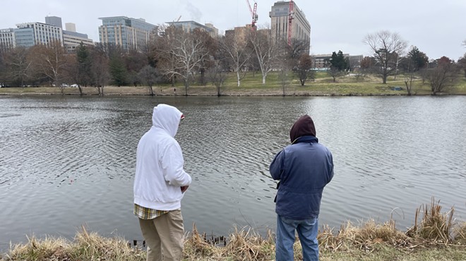 Two men stand in front of a lake in Forest Park with their backs turned to the camera. The man to the left wears a white hoodie and the man to the left wears a blue jacket.