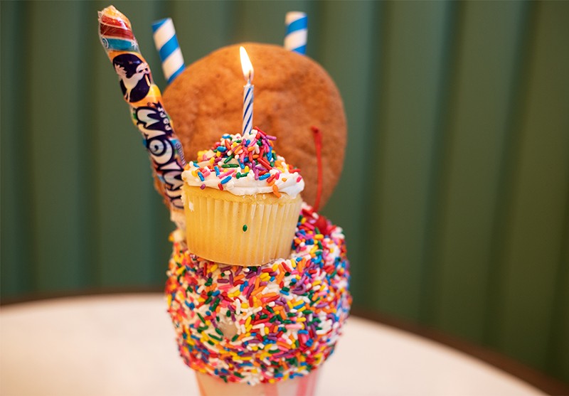 A Very Happy Un-Birthday Freak Shake with a vanilla-frosted rim, rainbow sprinkles, a funfetti cupcake, whipped cream, a snickerdoodle cookie, a cherry and a rainbow lollipop. - MABEL SUEN