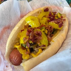 The St. Louis-Style hot dog is on a Vitale's roll lined with provolone cheese. The smoked and grilled all-beef hot dog is topped with grilled onions, green peppers, banana peppers, bacon and Steve's housemade smoky pepper mustard. - VIA STEVE'S HOT DOGS AND BURGERS