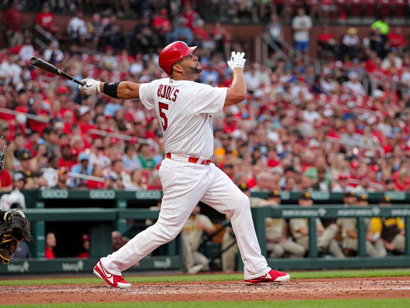 Albert Pujols, dressed in an all-white Cardinals uniform, finishes his swing as fans watch in the background.