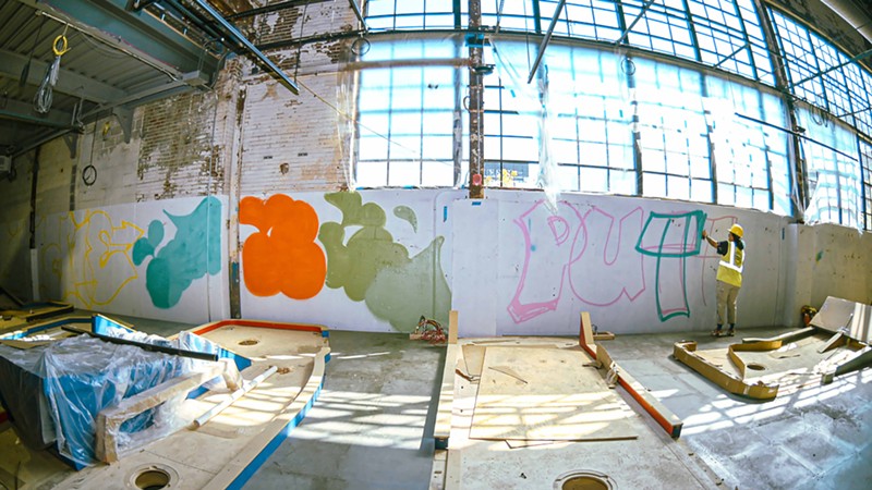 The very start of a mural on a blank wall takes shape beneath Indie 184’s spray cans.
