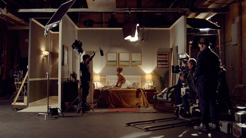 A film crew films and lights a woman who is sitting on a bed, partially unclothed.