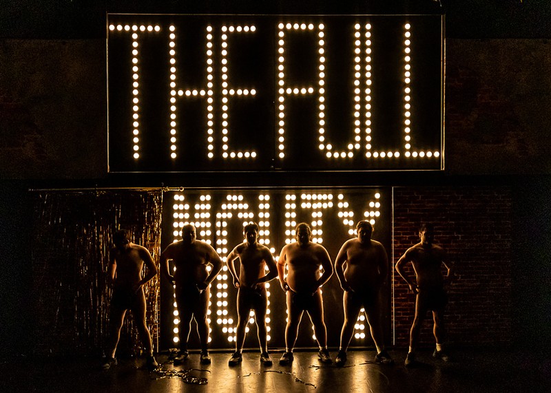 A group of men who are mostly unclothed stand on a stage in front of lights that spell out The Full Monty.
