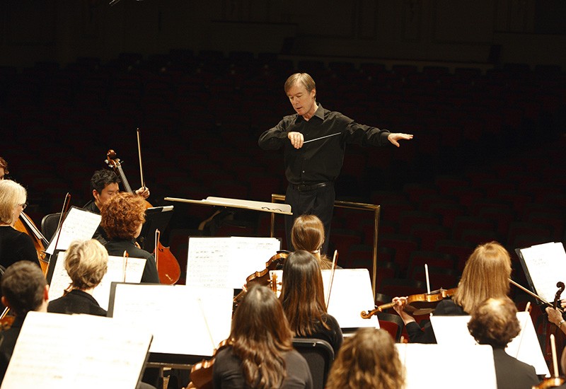 A conductor stands in front of a group of violinists.