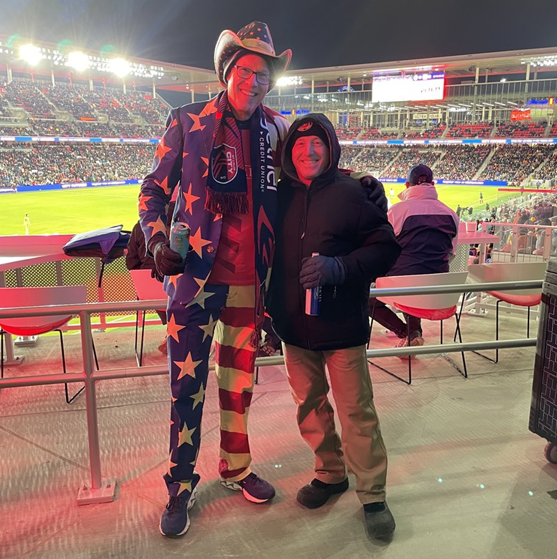 Two men pose for the camera in front of a brightly lit soccer stadium. The person on the left wears a suit and hat in the theme of the American fleg. He also has a scarf dedicated to St. Louis CITY. He puts his arm of a man who is wearing a black jacket and tan pants.