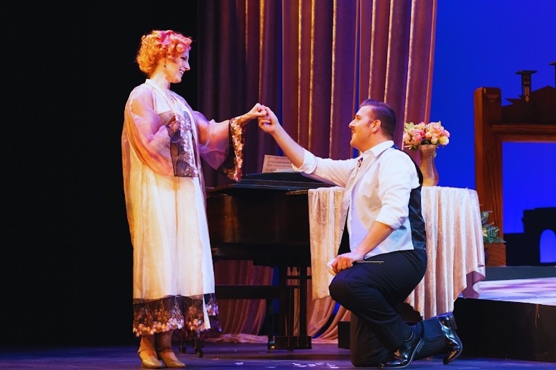 A woman dressed in 1920’s flapper style with a flower in her hair stands as a man dressed in trousers, vest and a shirt, kneels before her offering her praise, in a scene from Puccini’s opera ‘La Rondine’ at Winter Opera Saint Louis.