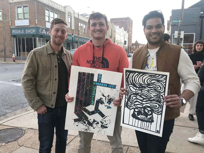 Three men stand together and two hold up prints from the Print Bazaar.