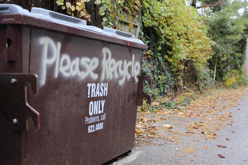 The words "Please Recycle" are spray painted above a brown dumpster that reads "Trash Only."