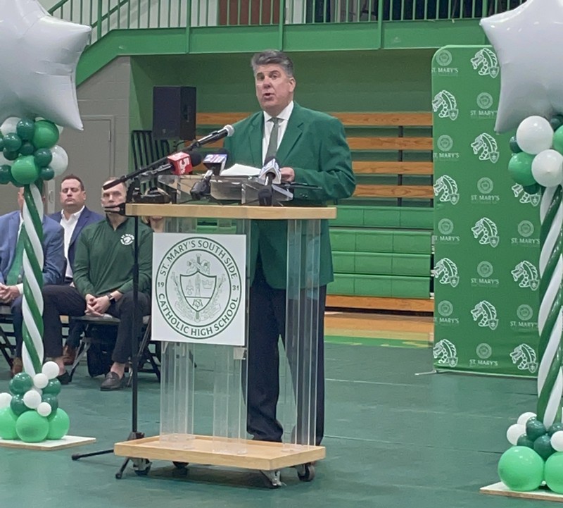 Mike England wears a green suit as he stands at a podium on a green floor in front of a green St. Mary's background.