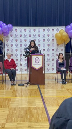 Cynthia Goudy stands at a podium at a press conference with purple and yellow ballons on either side.