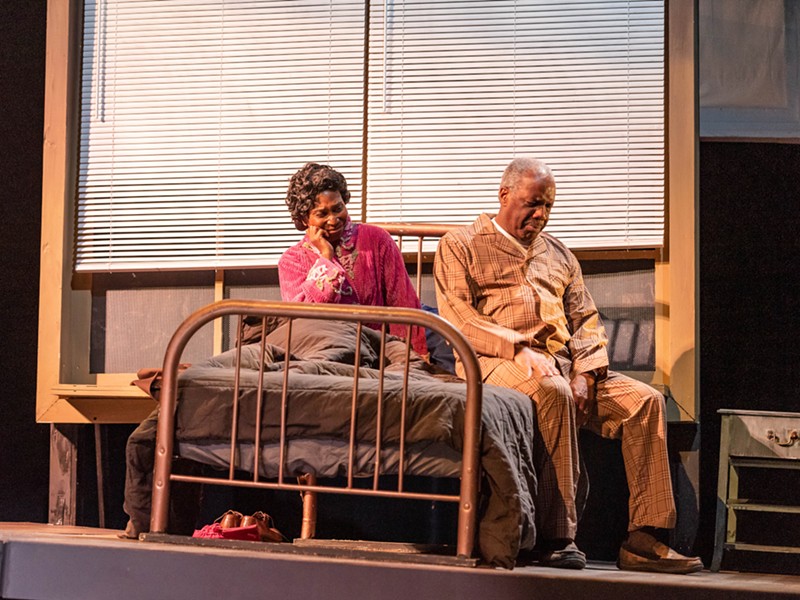 Two actors sit on a bed on stage, their faces contorted with emotion.