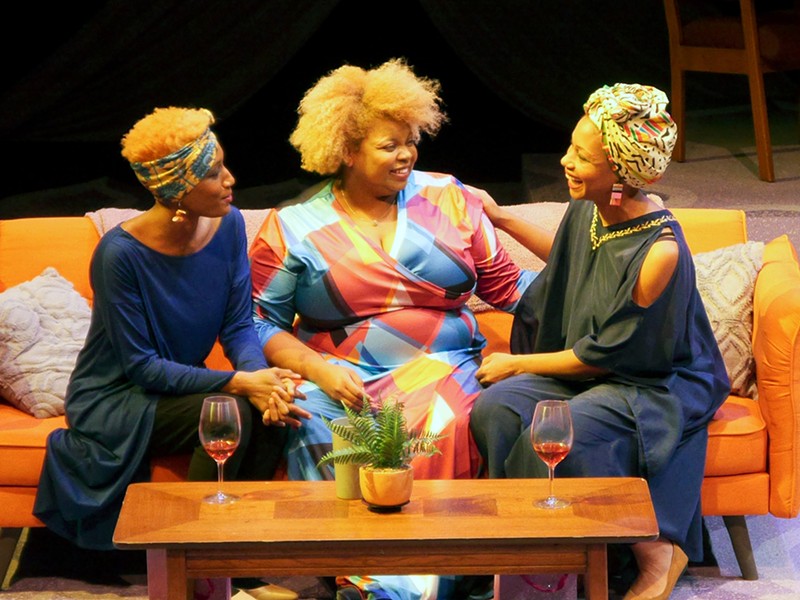 Three women sit on a couch on stage.