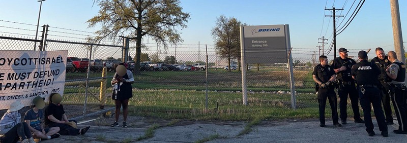 Protestors sit in front of the gate to Boeing's St. Charles plant while police huddle. - COURTESY ST. LOUIS FOR PALESTINE